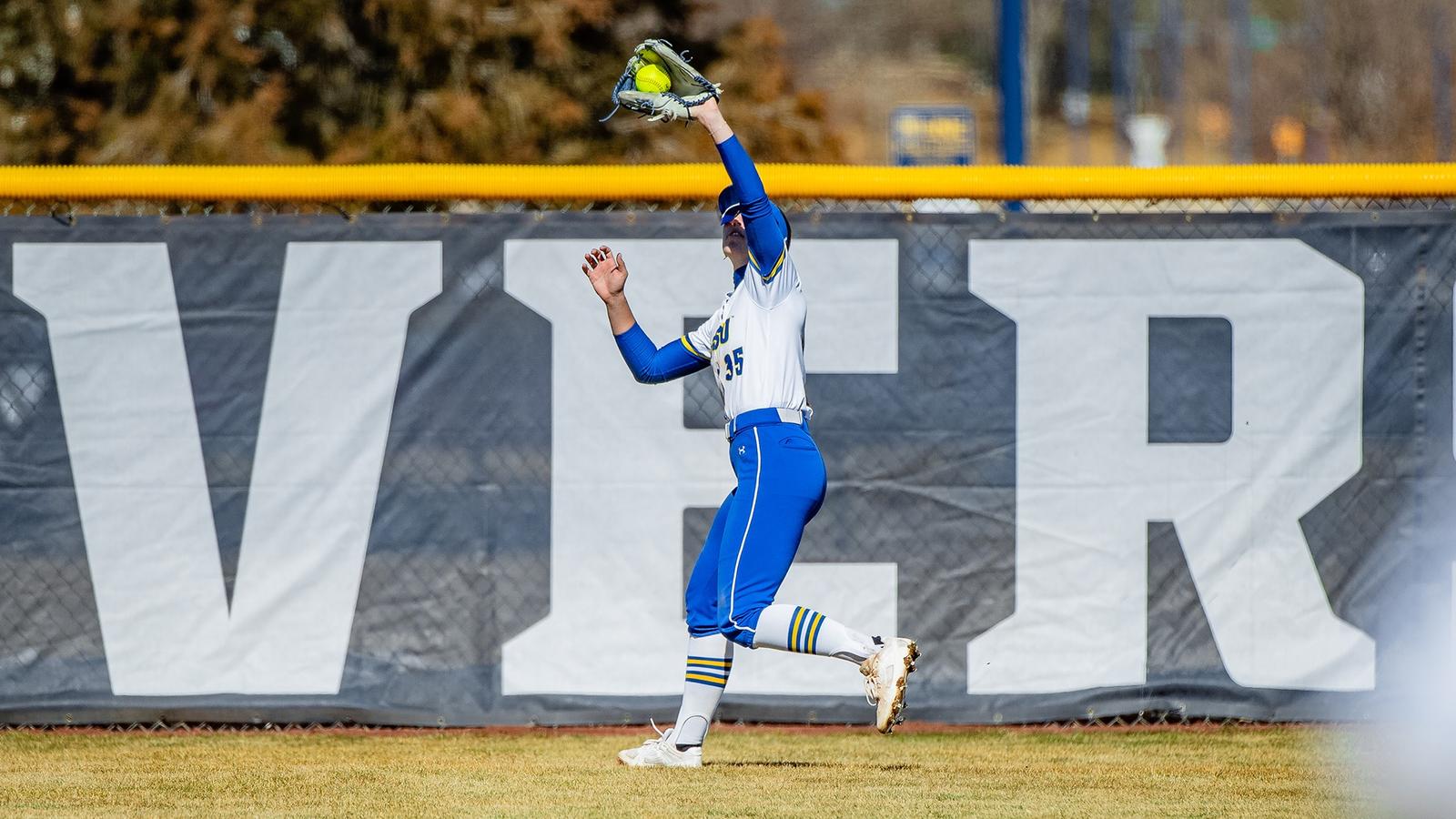 A softball player wearing an SDSU uniform and blue helmet stands on the field holding a ball, with the outfield fence and a building visible in the background.
