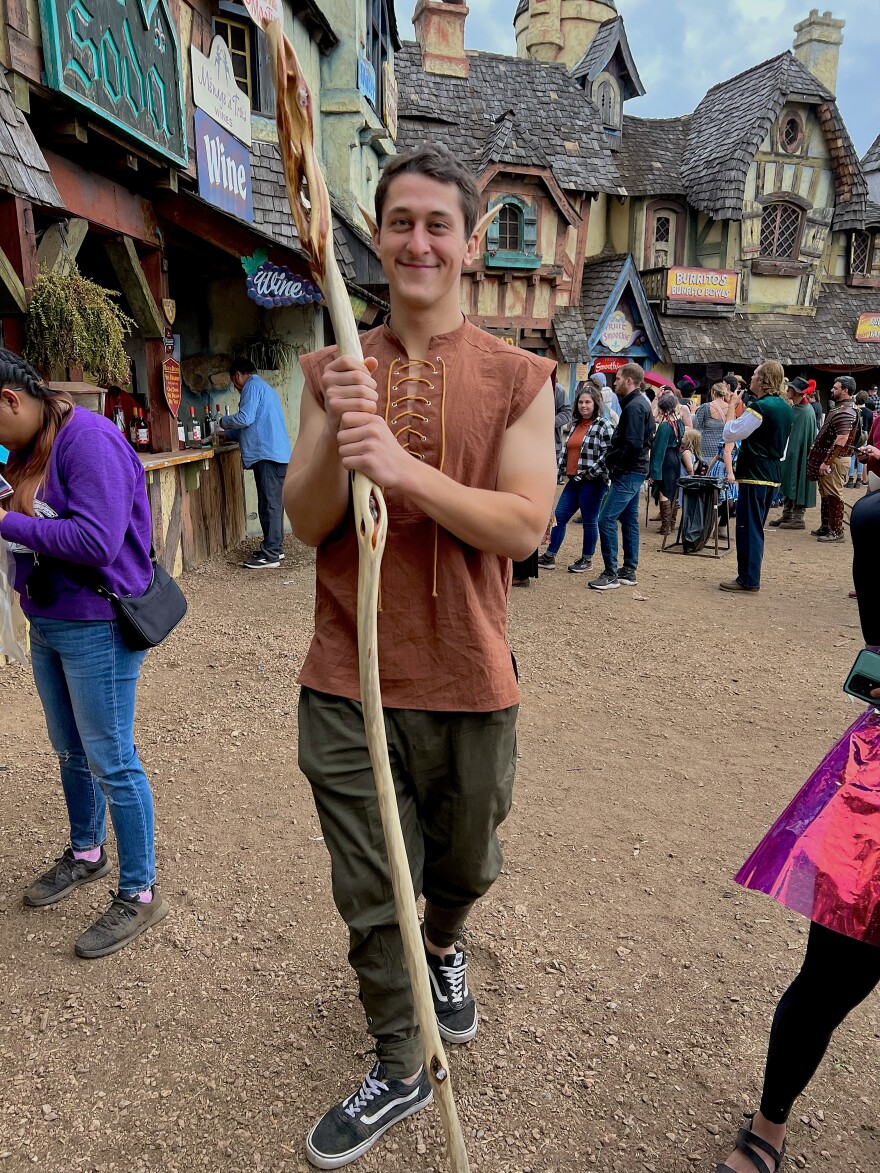 A man holding a wooden staff at a renaissance festival smiling for the camera.
