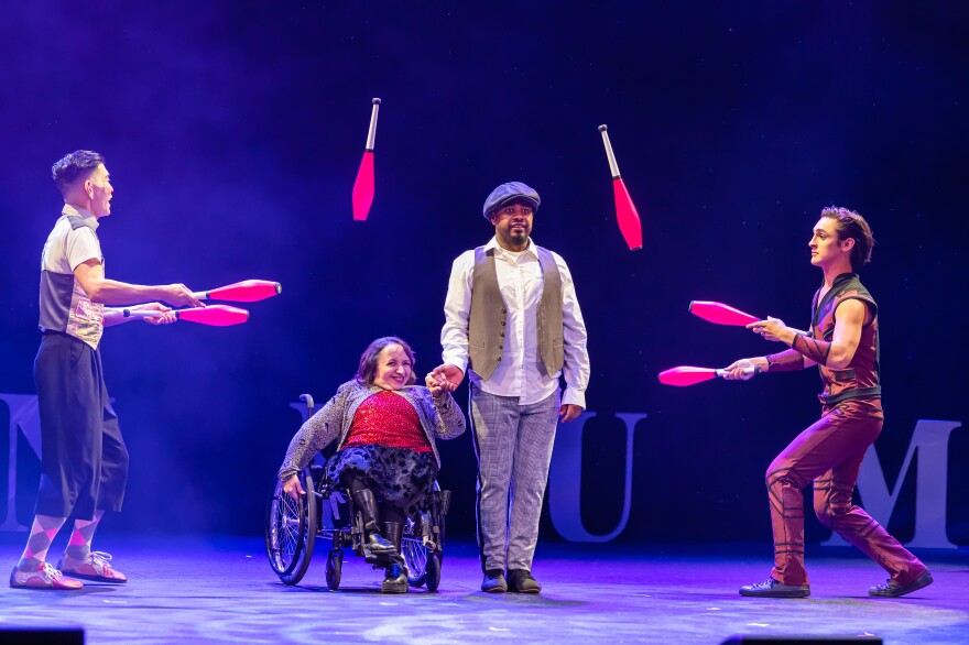 Two men juggle, passing the red juggling clubs past two other people in the middle, one standing and one seated in a wheelchair.