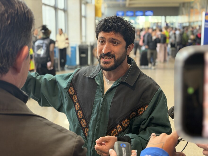 A man in a green jacket speaks to reporters inside the Barbara Jordan Terminal. Travelers pulling luggage can be seen behind him. 