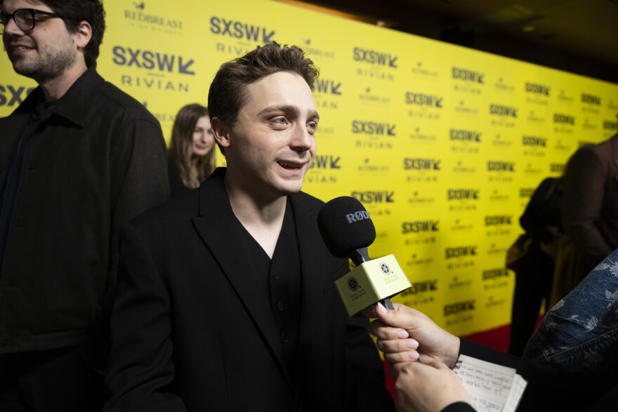 Jake Shane speaks into a microphone held by a member of the press while standing in front of a yellow backdrop with SXSW logos.