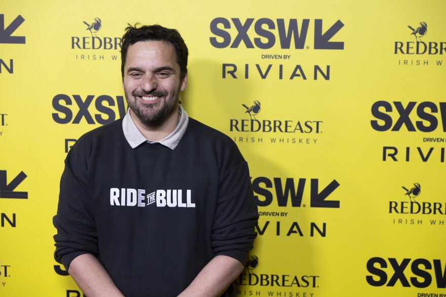 Jake Johnson smiles in front of a yellow backdrop with SXSW logos on it.