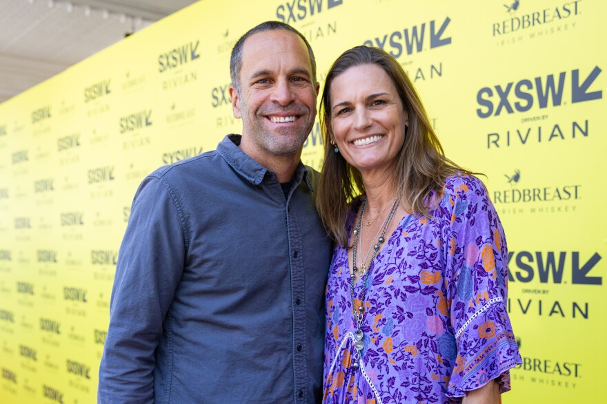 Jack Johnson and Kim Johnson stand, smiling, in front of a yellow backdrop with SXSW logos on it.