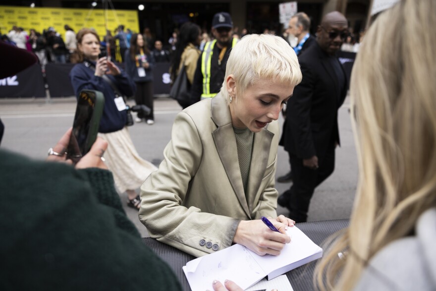 Emma Chamberlain signs an autograph while walking down a line of fans.