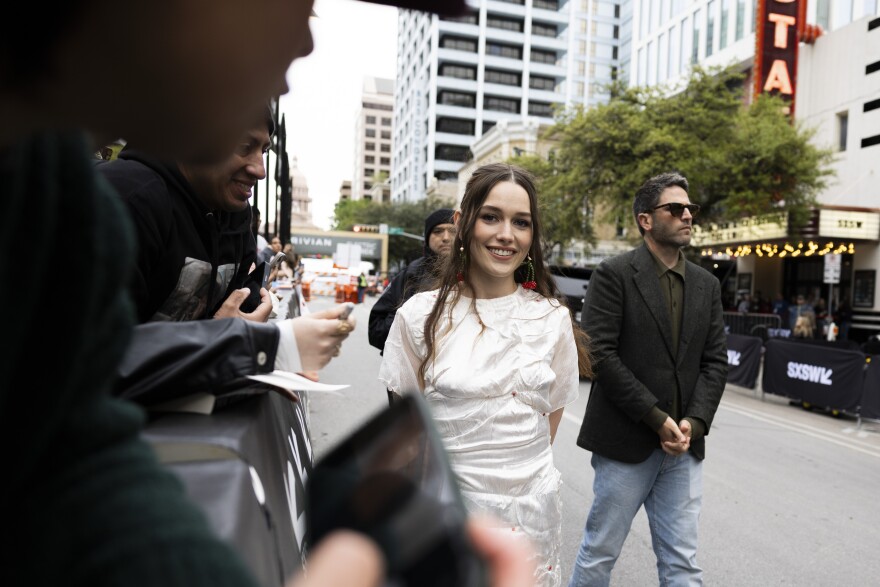 Victoria Pedretti smiles at the camera while walking down barricade lined with fans.