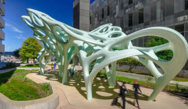 Outdoor public art installation featuring a large, pale green sculptural canopy with flowing, organic forms and cutout openings. The structure is supported by multiple curved legs and casts patterned shadows on the ground below. People walk beneath the sculpture along a landscaped pathway, with modern buildings and trees visible in the background under a clear blue sky.