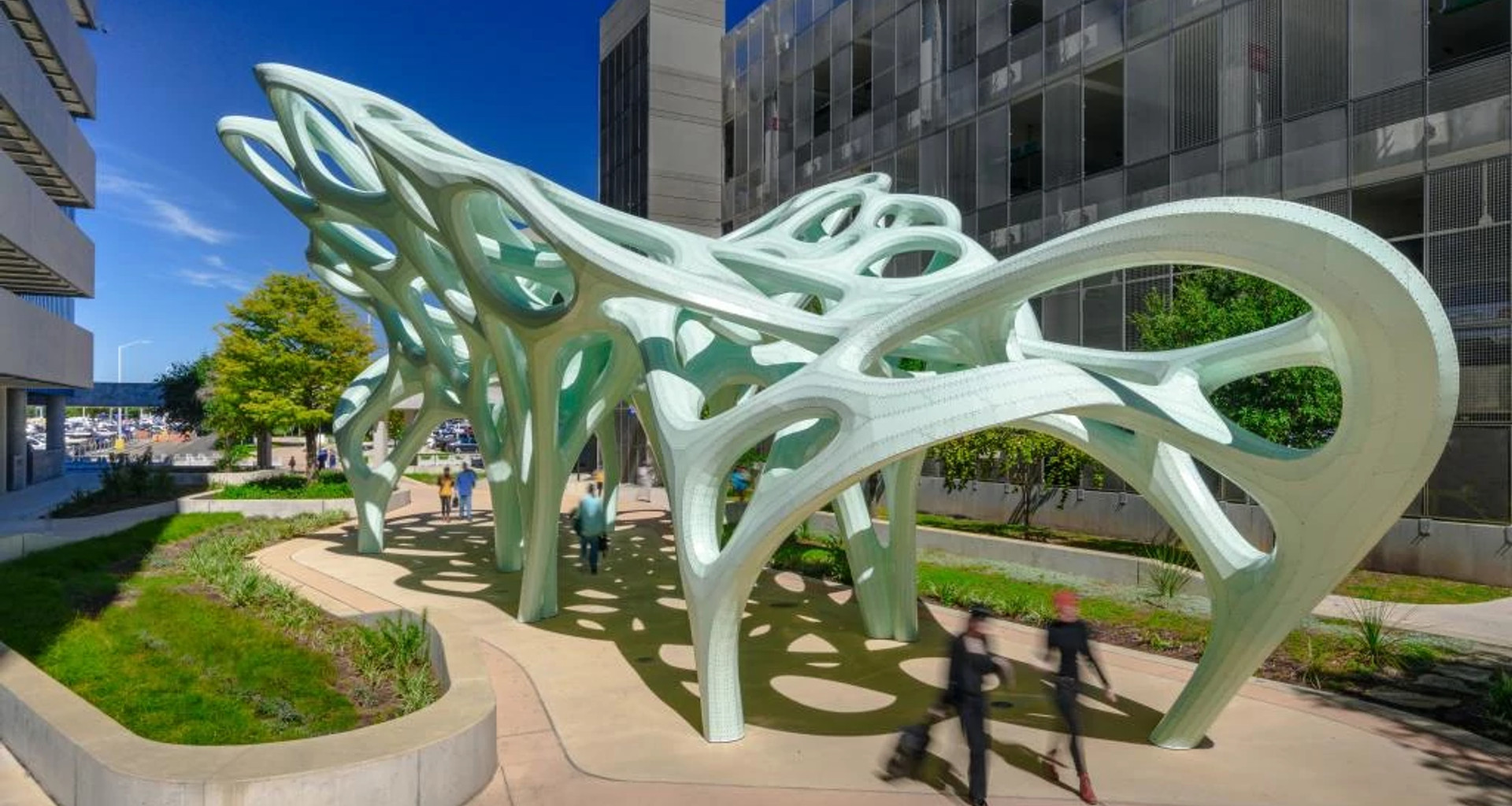 Outdoor public art installation featuring a large, pale green sculptural canopy with flowing, organic forms and cutout openings. The structure is supported by multiple curved legs and casts patterned shadows on the ground below. People walk beneath the sculpture along a landscaped pathway, with modern buildings and trees visible in the background under a clear blue sky.