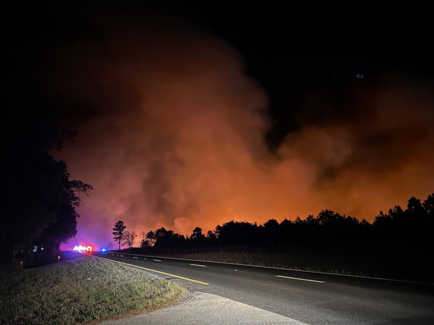 Fire, smoke and emergency vehicles can be seen in the distance past a road in a rural area. 