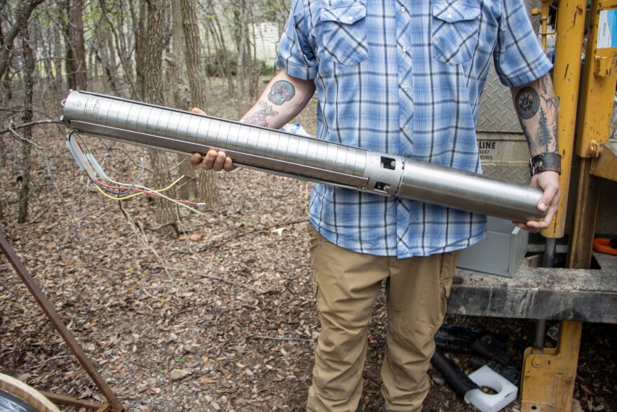 A man standing outside amongst trees holds a long, silver, pipe.