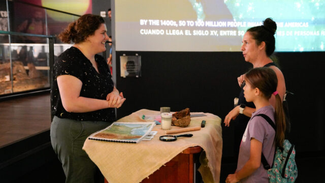 A woman standing at a table with touchable objects talking to two museum visitors.