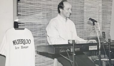 A black and white image of Musician Floyd Domino playing a keyboard next to a Waterloo Ice House t-shirt display.