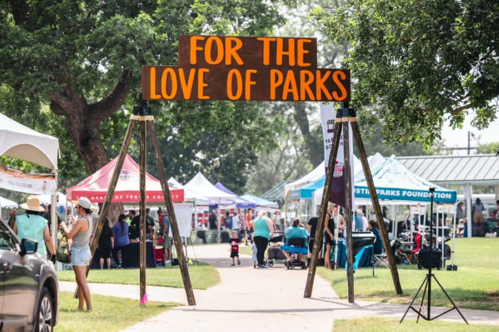 This image depicts a wooden event sign that reads, "For the Love of Parks," at Richard Moya Park. There are several tents in the background featuring different businesses and organizations.