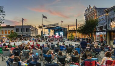 A large crowd fills a blocked-off downtown street at dusk, seated in lawn chairs facing a large outdoor stage flanked by historic brick storefronts.