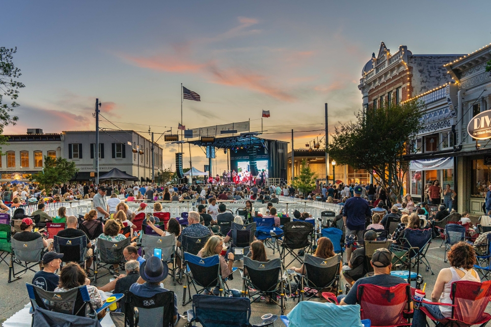 A large crowd fills a blocked-off downtown street at dusk, seated in lawn chairs facing a large outdoor stage flanked by historic brick storefronts.