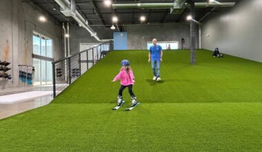 Interior of a business featuring a manmade hill covered in astroturf with a young female wearing skis coming down the slope and a male walking behind her.