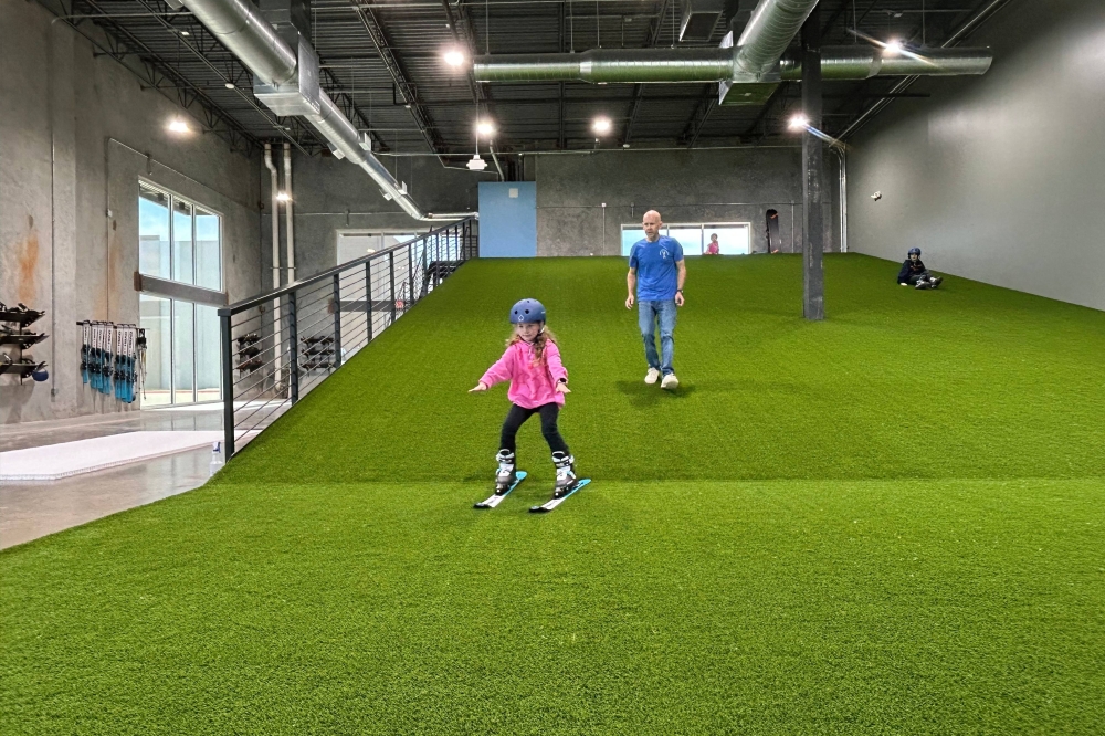 Interior of a business featuring a manmade hill covered in astroturf with a young female wearing skis coming down the slope and a male walking behind her.