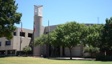 The exterior of the Plano Municipal Center, a blocky gray building, partially obscured by trees
