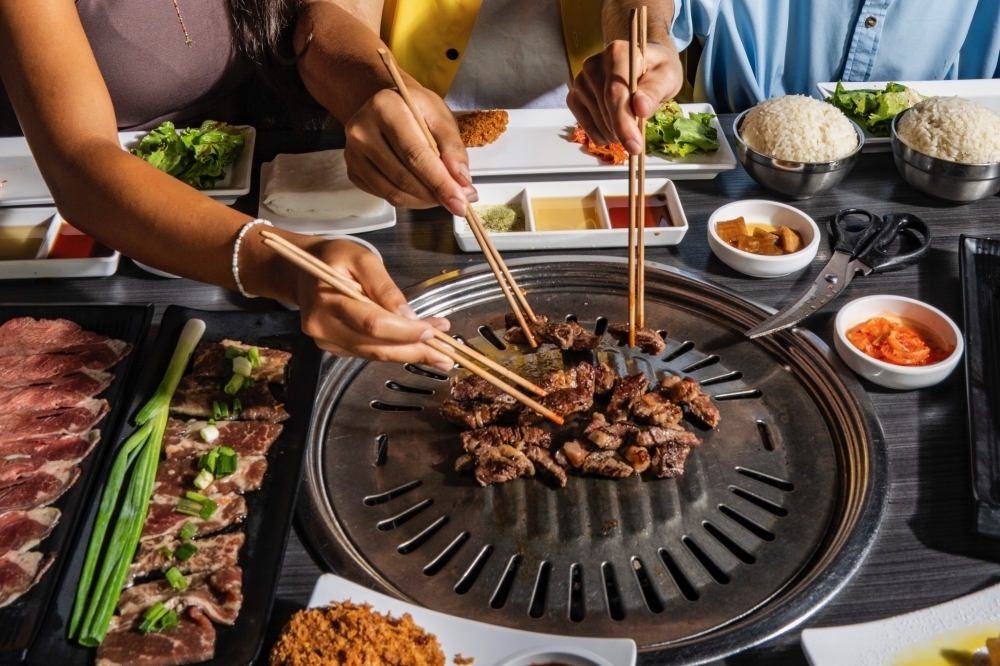 Overview of people's hands cooking strips of meat on a table-top grill.