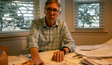 James Dodson is looking at the camera with a serious expression. On the table in front of him are maps and documents. Behind him are windows, the shades open, trees beyond them.