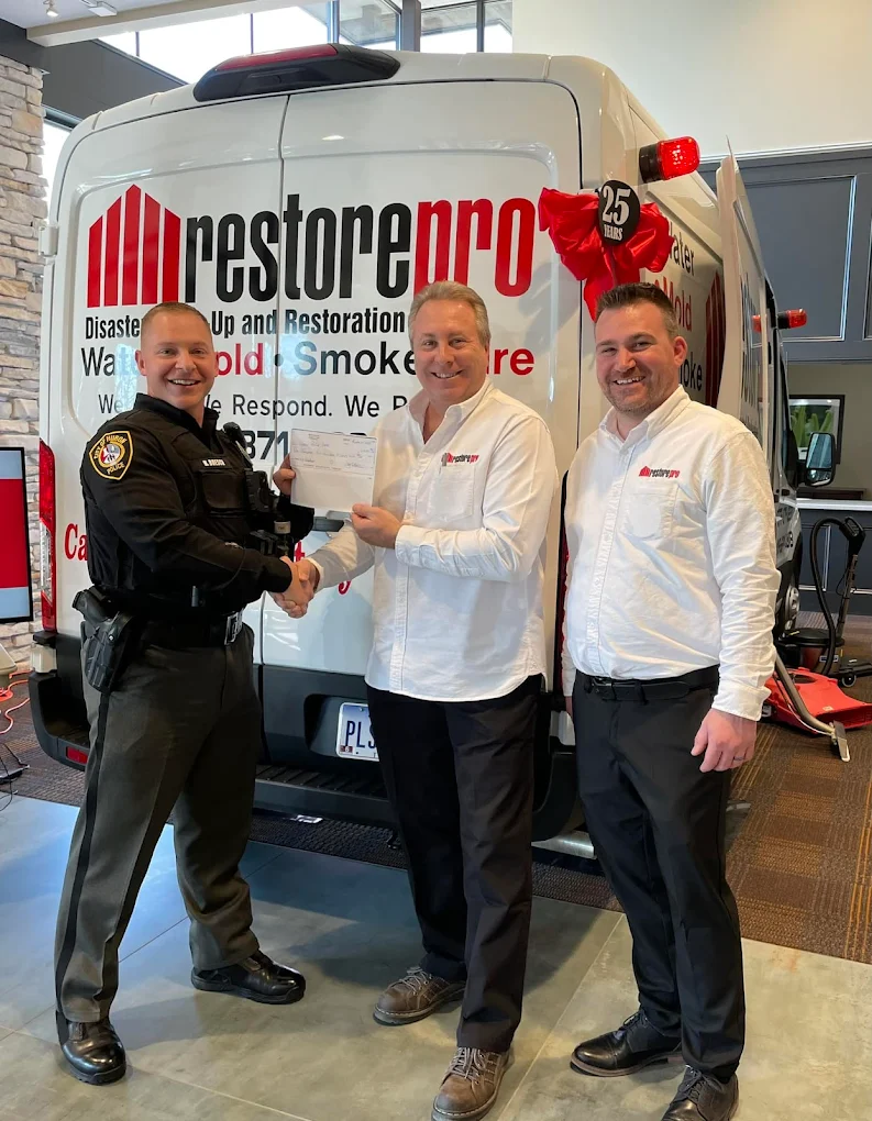 RestorePro team members pose with a uniformed officer in front of a disaster restoration van during a check presentation.