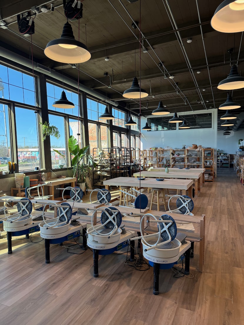 Rows of electric pottery wheels and prep tables fill the airy studio, while pieces in...