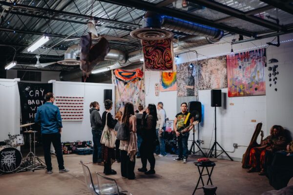 People gathered in an artist studio with paintingsandbanners on the walls, music equipment, and a musician with a guitar.