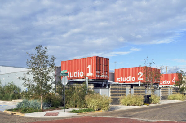 A one-story modern building with orange containers suspended above multiple entrances, each labeled "studio 1, 2, 3, 4."