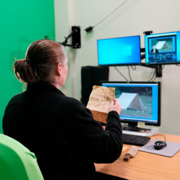 A woman holding up an aged piece of paper in front of three monitors.