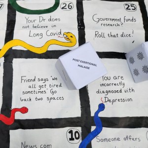 A close up of the board game All Snakes No Ladders features a six-sided die with little viral blobs as dots. The spaces read: "Your doctor does not believe in Long COVID." "Government funds research? Roll that dice!" "Friend says 'We all get tired sometimes.' Go back two spaces." "You are incorrectly diagnosed with depression."