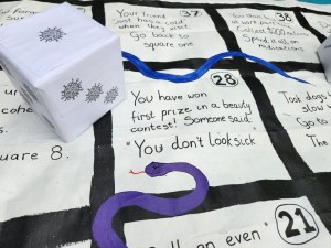 A close up of the board game All Snakes No Ladders features a six-sided die with little viral blobs as dots. The space in the middle reads, "You have won first prize in a beauty contest. Someone said, 'You don't look sick.'"