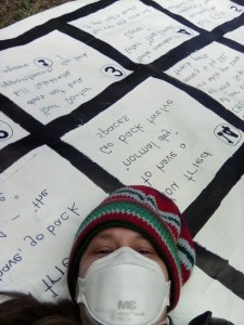 A person with Long COVID from the Australian Long Covid Community wears a respirator and lies down on top of a giant board game called All Snakes No Ladders.
