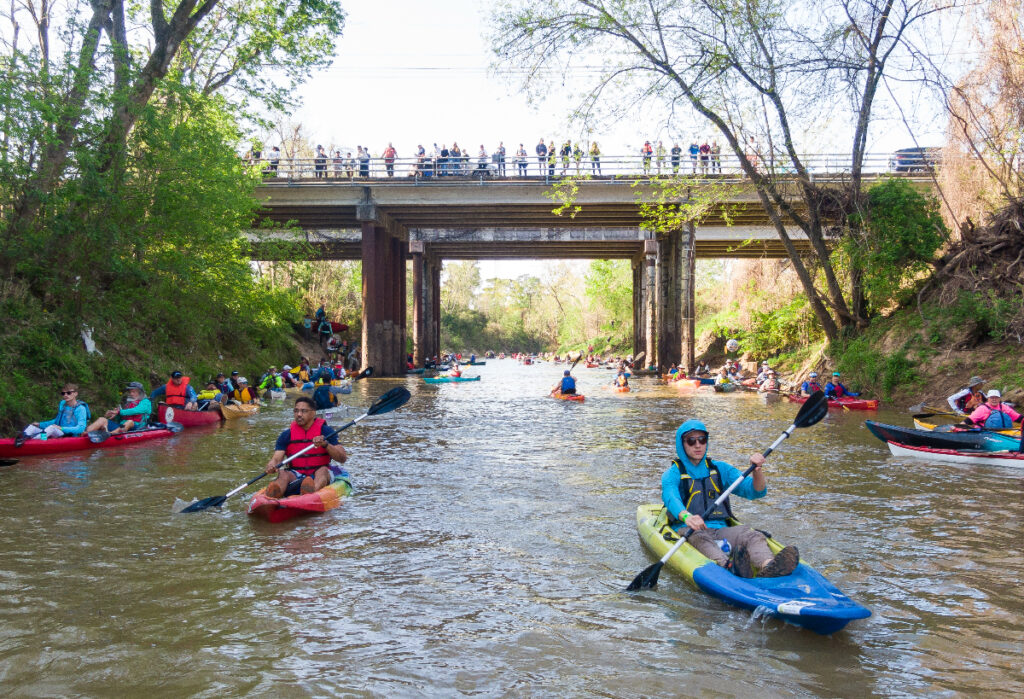 Image shows paddlers in kayaks and canoes riding down Buffalo Bayou in Houston.