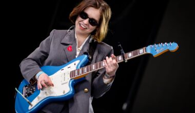 in a gray suit and black sunglasses, Lindsey Jordan of snail Mail performs on stage with a blue and white guitar