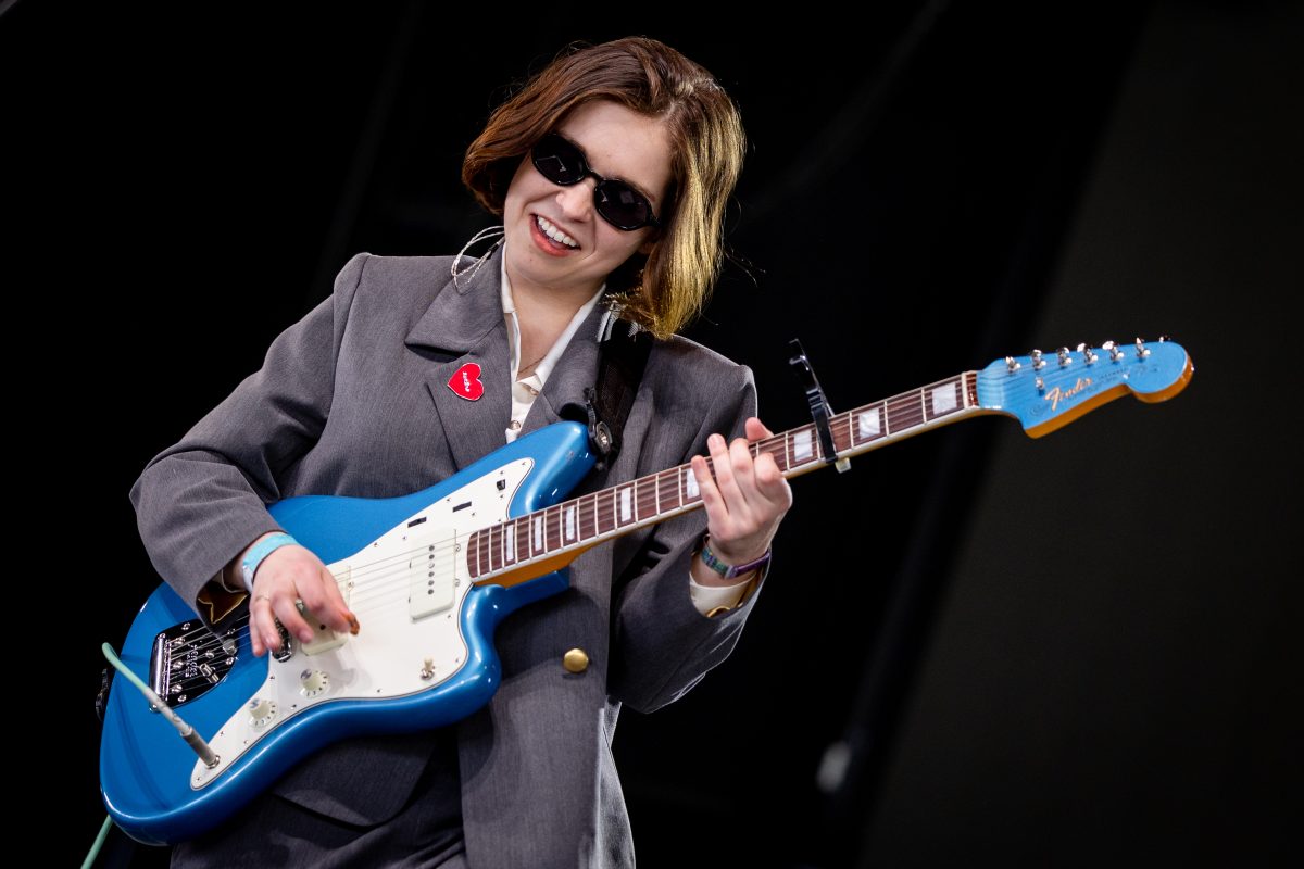 in a gray suit and black sunglasses, Lindsey Jordan of snail Mail performs on stage with a blue and white guitar