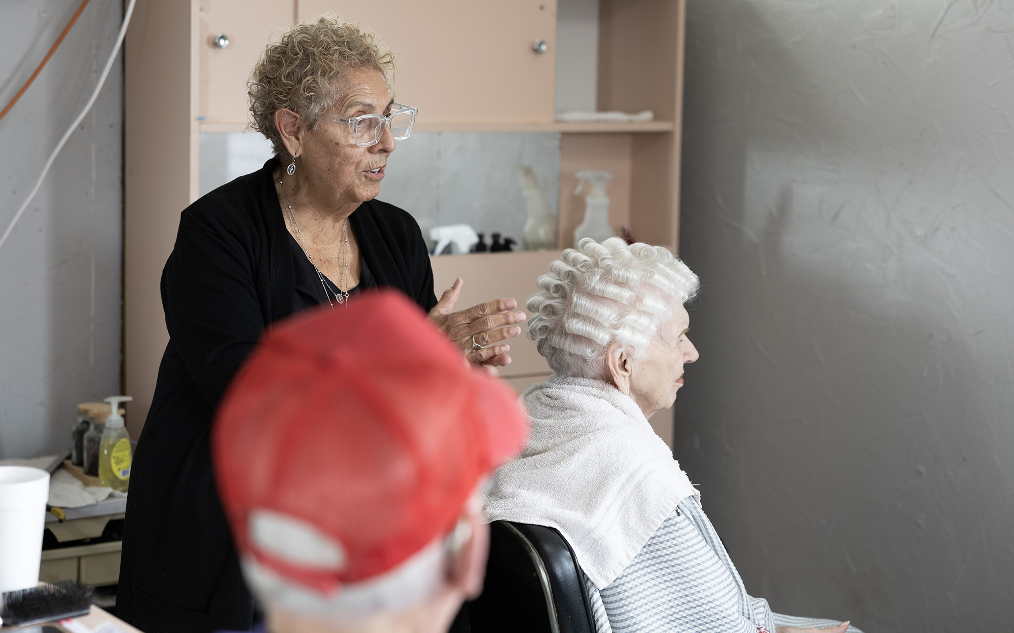 A woman works on the hair of another elderly woman, seated with a towel around her shoulders, while a man in a red baseball cap looks on.