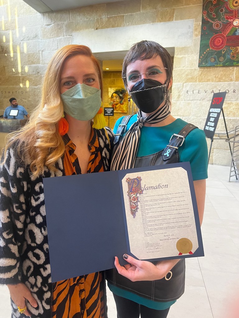 Two Long COVID advocates wearing respirators stand side-by-side holding the city of Austin’s Long Covid Awareness Day Proclamation.