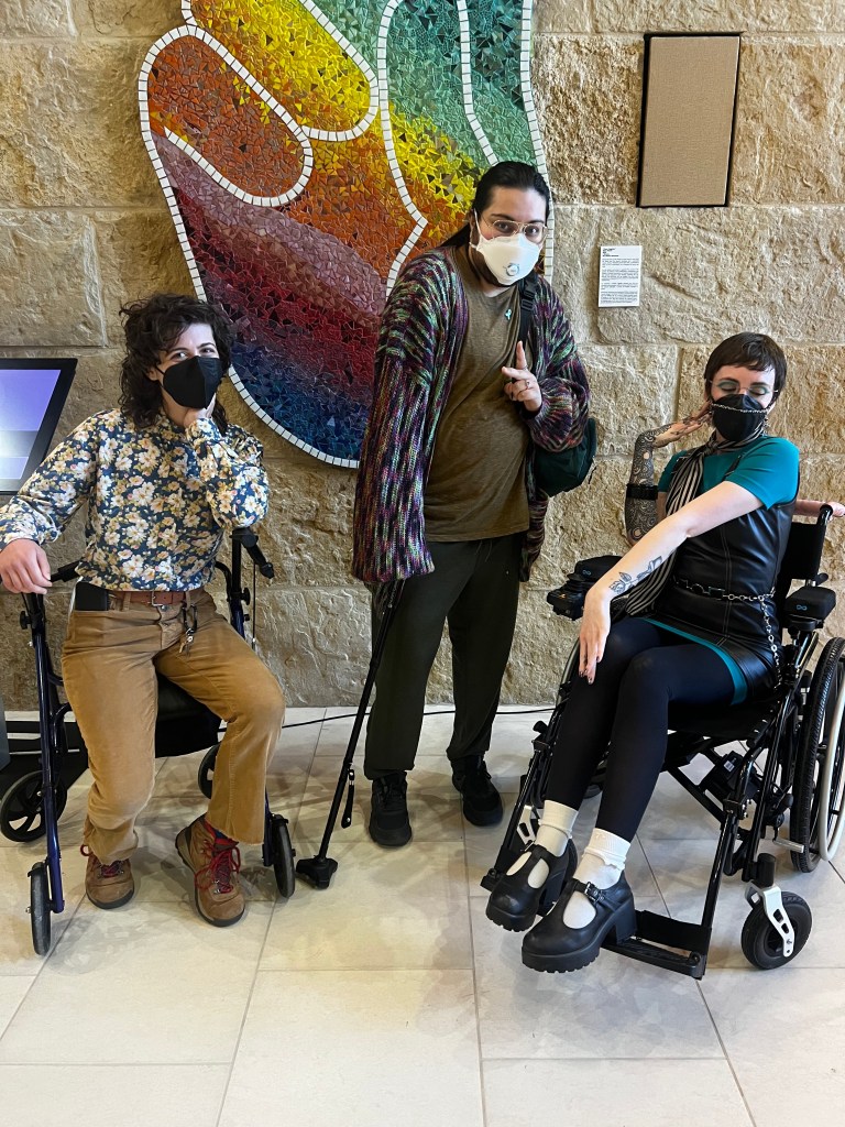 Three Long COVID advocates in Austin stand in front of a rainbow-colored peace sign mural. All three are wearing respirators and using different mobility aids: a rollator, a cane, and a wheelchair.