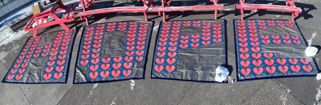 An art display of red broken hearts against blue backdrops, laid out on a black tarp. On the left side of each heart, the text reads, “Having Long COVID is heart-breaking because…” The right side of each hearts lists a reason Long COVID is heart-breaking with an attribution for the person with Long COVID who shared their thoughts.