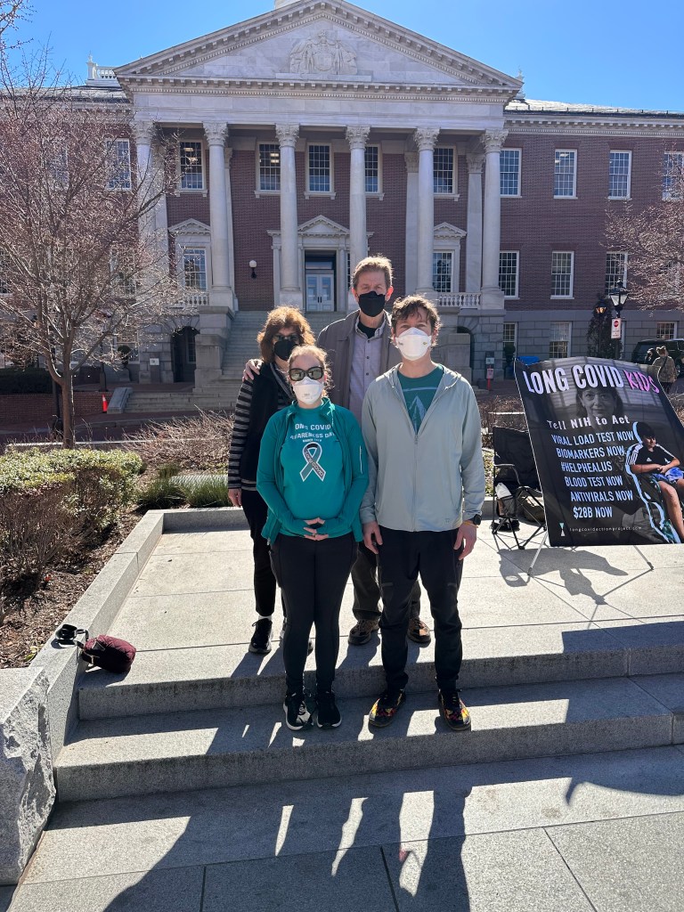 A small group of advocates wearing respirators stands in front of the Maryland State House beside a poster that reads “Long COVID Kids.”