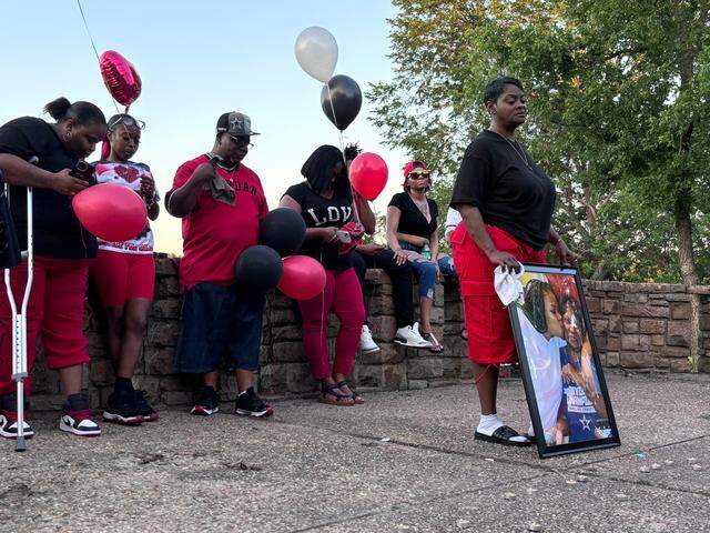 A woman in a black shirt and red pants holds a large photo of herself with a younger woman. She has a downcast look on her face. Behind her, others dressed in red and black sit on a low stone wall holding red, black and white ballons.