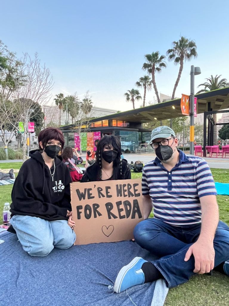 Three people in respirators sit outside on a blanket holding a sign that says, "We're here for Reda."