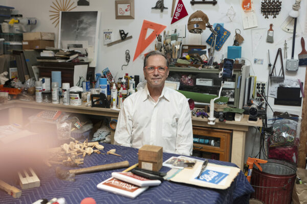 A photograph of artist Jack Massing sitting at a work table in his studio with a blurred hand in the foreground.