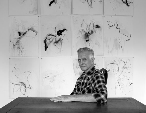 A black and white photograph of artist Joseph Havel sitting at a table with an array of loose ink paintings of flowers on a grid of paper behind him.