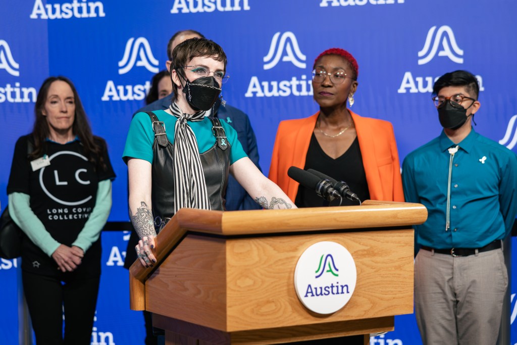 Katie Drackert delivers a speech at the Austin city council meeting where councilmembers recognized International Long COVID Awareness Day, while fellow advocates look on in the background. Katie is wearing a black respirator with a mask chain.