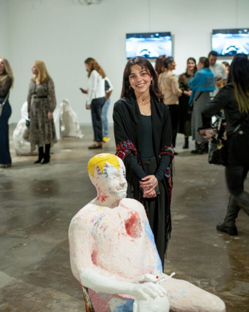 A young artist stands with her figural sculpture in a gallery space with other people looking at artworks.