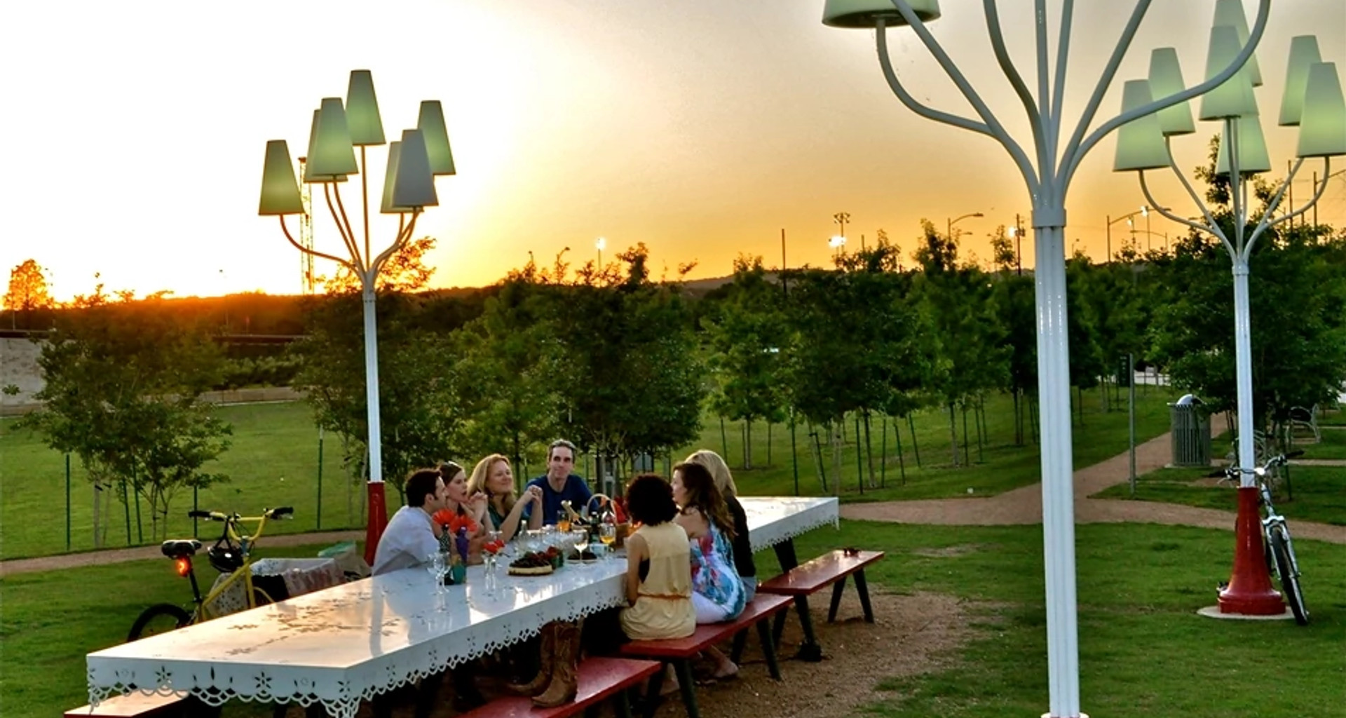 Six people gathered around a long white outdoor table with red benches in a park at sunset, enjoying food and drinks beneath whimsical tree-like lamp posts, with bicycles nearby and a serene landscape of grass, paths, and young trees in the background.