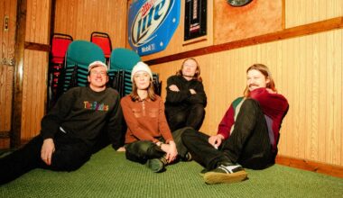 indie rock band ratboys sits on green carpet surrounded by wood-paneled walls with a blue miller lite sign and dart board hanging above