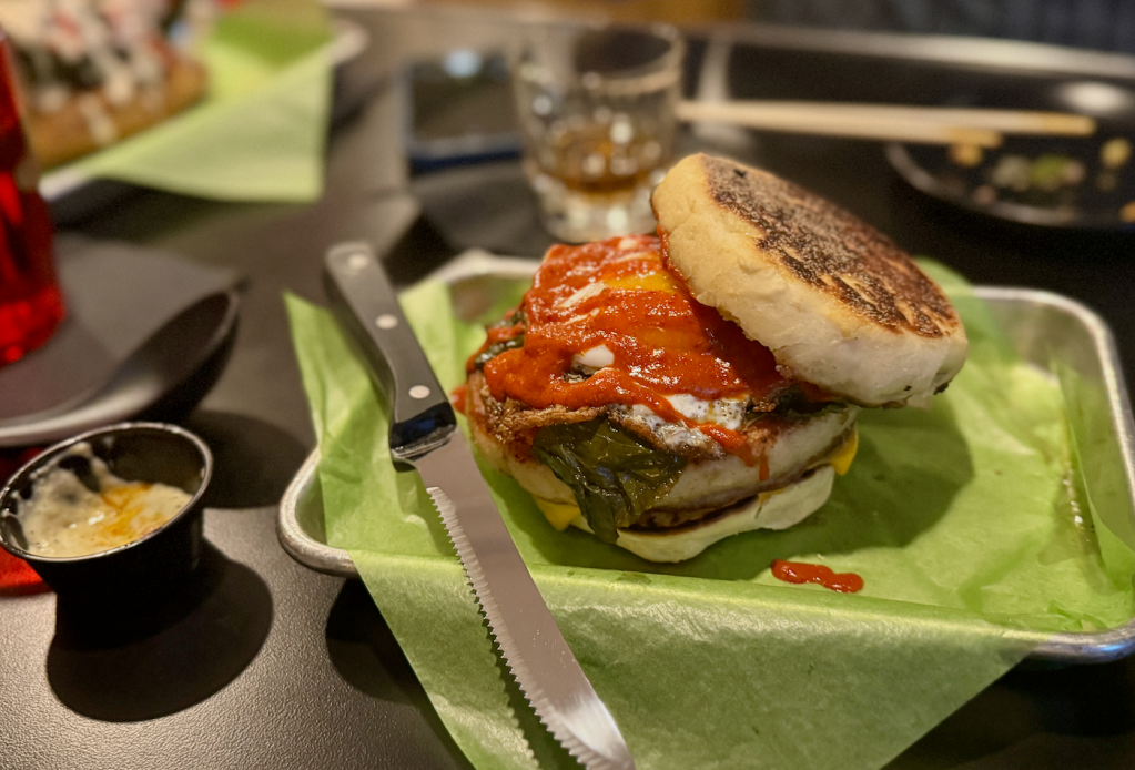 A close up of Rainbowcat's McMuffin that takes a house-made English muffins stuffed with kasu-cured porchetta, hot sauce, braised greens, fried egg and pickled radish.