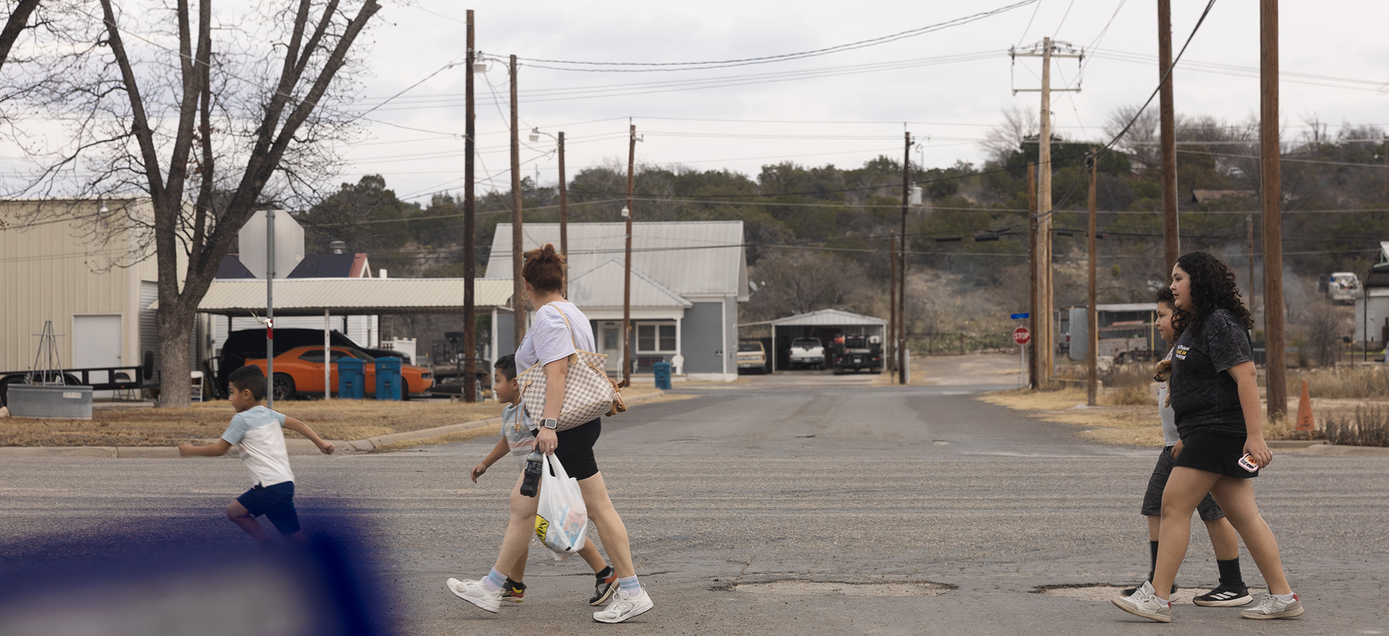 A woman holding a grocery bag and four children cross a quiet street