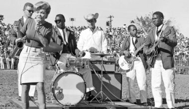 A black-and-white photo showing a White woman with a bouffant hairstyle singing into a microphone on the left side of the frame on the dirt grounds of a rodeo, while in the background are Black and White incarcerated men, wearing dark colored jackets, black ties and billowing white pants playing guitars and a drum. The drummer is wearing sunglasses, as a correctional officer stands guard behind him dressed in white and a cowboy hat with the brim upturned on both sides. In the distance, crowds sit in the stands.